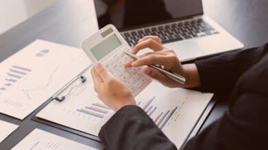 Person calculating minimum salary for a car loan, with financial charts and laptop on the desk.
