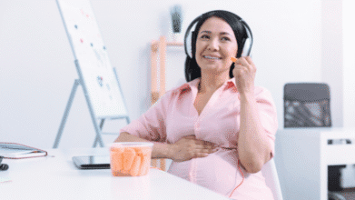 Smiling woman listening to music and snacking at her desk for a relaxing Mother's Day 2025 moment