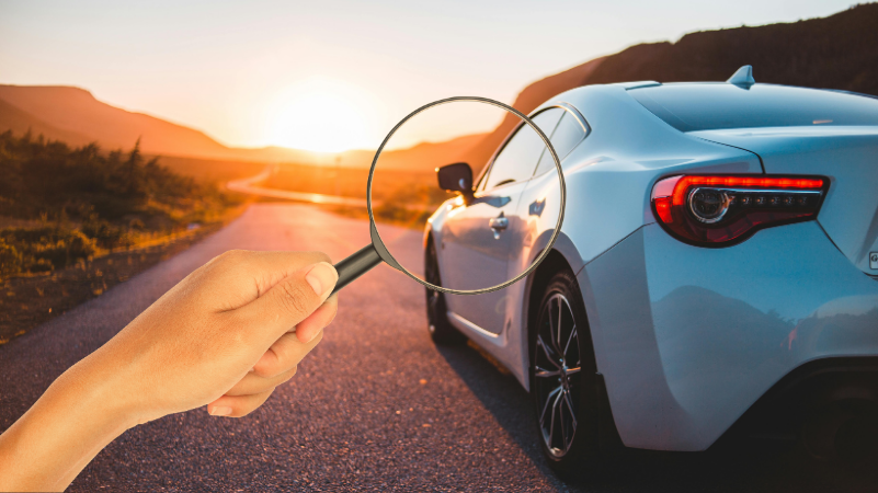 Hand holding magnifying glass inspecting a car, symbolizing research before buying a secondhand car.